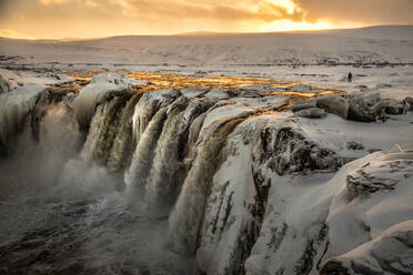 Majestätischer Blick auf einen reißenden Wasserfall, der im Winter in Island bei Sonnenuntergang einen gefrorenen Felsen hinunterfließt - ADSF37398