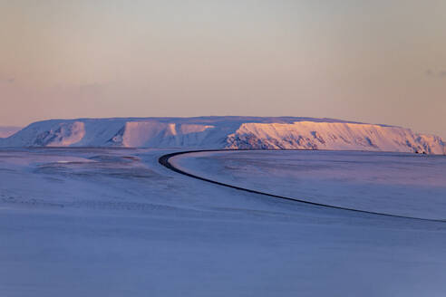 Leere, kurvenreiche Asphaltstraße, die durch ein schneebedecktes Tal im Hochland führt, bei Sonnenuntergang im Winter in Island - ADSF37399