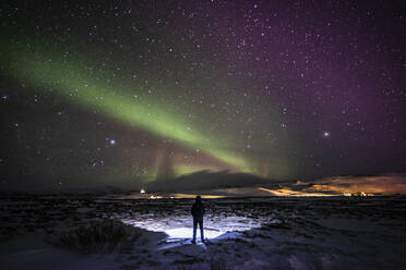 Silhouette of unrecognizable traveler standing on snowy terrain and enjoying view of green polar lights glowing in night starry sky in Iceland - ADSF37401