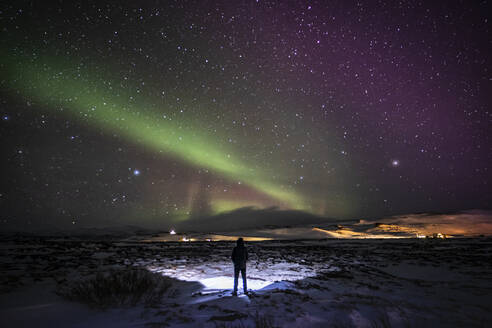 Silhouette eines nicht erkennbaren Reisenden, der auf einem verschneiten Gelände steht und die Aussicht auf das grüne Polarlicht am nächtlichen Sternenhimmel in Island genießt - ADSF37401