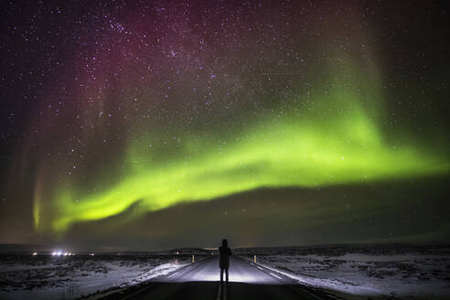 Silhouette eines nicht erkennbaren Reisenden, der auf einer asphaltierten Fahrbahn steht und den Blick auf die grünen Polarlichter am nächtlichen Sternenhimmel in Island genießt - ADSF37402