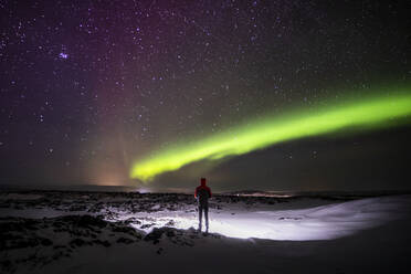 Silhouette eines nicht erkennbaren Reisenden, der auf einem verschneiten Gelände steht und die Aussicht auf das grüne Polarlicht am nächtlichen Sternenhimmel in Island genießt - ADSF37403