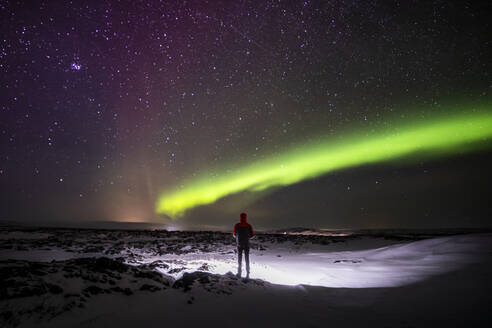 Silhouette of unrecognizable traveler standing on snowy terrain and enjoying view of green polar lights glowing in night starry sky in Iceland - ADSF37403