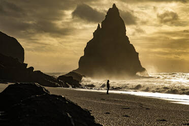 Unrecognizable traveler silhouette in warm clothes and hat walking admiring dramatic landscape of volcanic coastline black sand beach with rock formations washed by foamy waves of stormy sea in Iceland - ADSF37428