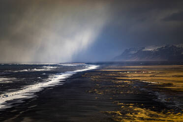 Vulkanische Küste mit schwarzem Sand, umspült von schäumenden Wellen des stürmischen Meeres, mit einem Dorf in der Nähe der Berge in Island - ADSF37430