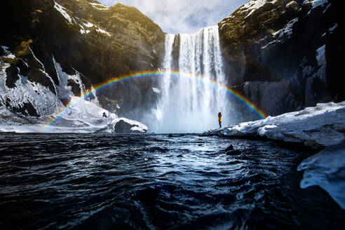 Unerkennbarer Reisender in der Nähe eines beeindruckenden Wasserfalls und eines Regenbogens in den Bergen während eines Urlaubs in Island - ADSF37431