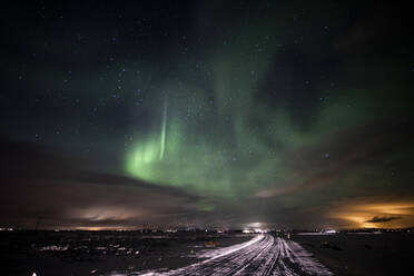 Gerade Straße, die durch verschneite Landschaft in Richtung Berge gegen den Himmel mit grünen Polarlichtern in der Winternacht in Island führt - ADSF37433