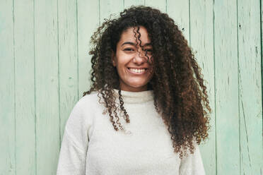 Happy young woman with curly hair standing in front of wooden wall - KIJF04499