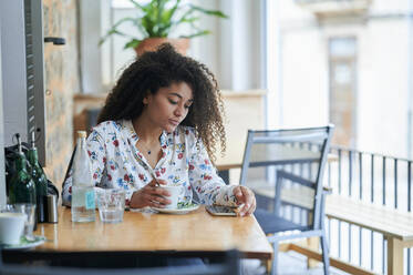 Junge Frau mit Kaffeetasse und Blick auf ihr Smartphone in einem Café - KIJF04502