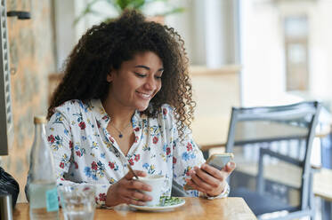 Happy young woman sitting with coffee cup and smart phone at cafe - KIJF04504