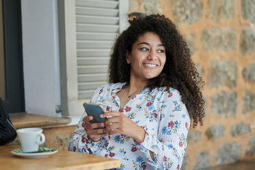 Thoughtful smiling young woman sitting with mobile phone at cafe - KIJF04505