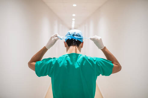 Back view of unrecognizable male doctor in green uniform and sterile gloves tying medical cap in narrow hospital hall - ADSF37505