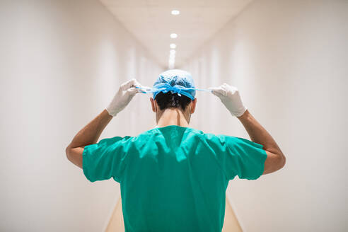Rückenansicht eines nicht erkennbaren männlichen Arztes in grüner Uniform und sterilen Handschuhen, der in einem schmalen Krankenhausflur eine medizinische Kappe bindet - ADSF37505
