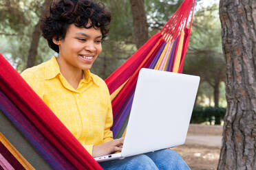 Lächelnde Teenagerin chillt in der Hängematte im Park und surft auf dem Laptop - ADSF37509