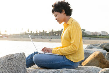 Seitenansicht einer jungen Frau in Freizeitkleidung, die auf einem Felsen am Meer sitzt und auf ihrem Laptop surft, während sie ihre Zeit am Meer verbringt - ADSF37517