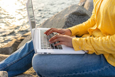 Ausgeschnittene, nicht erkennbare Frau in Freizeitkleidung, die auf einem Felsen am Meer sitzt und auf ihrem Laptop surft, während sie ihre Zeit am Meer verbringt - ADSF37518