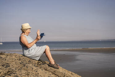 Smiling senior woman waving hand on video call sitting at beach - UUF27193