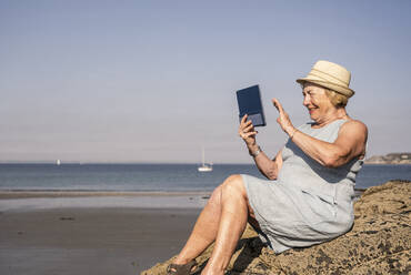 Glückliche ältere Frau mit winkender Hand bei einem Videoanruf am Strand sitzend - UUF27194