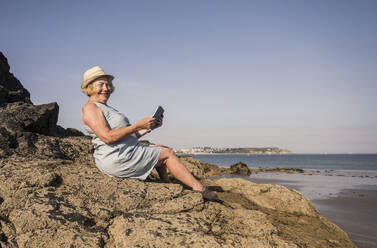 Happy woman with tablet PC sitting on rock at beach - UUF27198
