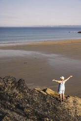 Ältere Frau mit erhobenen Armen auf einem Felsen am Strand stehend - UUF27201