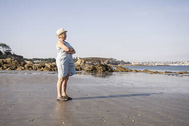 Ältere Frau mit verschränkten Armen am Strand stehend an einem sonnigen Tag - UUF27206