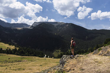 Junge Frau mit Rucksack steht vor einem Berg an einem sonnigen Tag - PNAF04504
