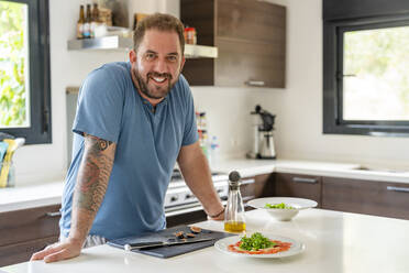 Happy mature man leaning on kitchen island at home - DLTSF03079