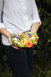 Mittelteil einer Frau, die eine Schüssel mit veganem Salat mit Gemüse und essbaren Blumen hält - EVGF04078