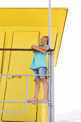 Lächelnder Junge mit verschränkten Armen, der auf einem Geländer vor einer gelben Rettungsschwimmerhütte steht - MEGF00006