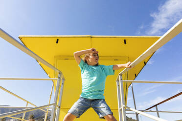 Boy shielding eyes standing amidst railings in front of yellow lifeguard hut - MEGF00027