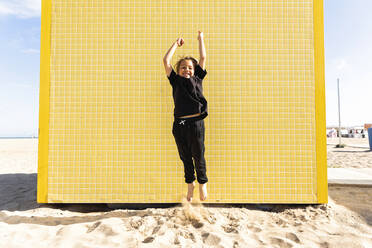 Cheerful boy jumping in front of yellow wall at beach - MEGF00031