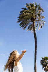 Girl with arm outstretched standing in front of palm tree on sunny day - MEGF00043