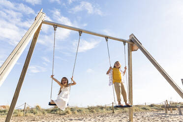 Cheerful girl with brother playing on swing at beach - MEGF00051