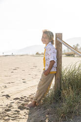 Lächelnder Junge mit Hand in der Tasche, der sich an einen Holzpfahl am Strand lehnt - MEGF00052