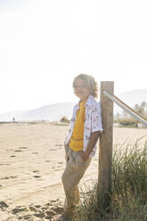 Lächelnder Junge, der an einem sonnigen Tag am Strand an einem Pfahl steht - MEGF00054
