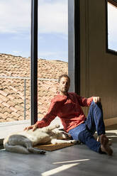 Man sitting with dog leaning on window at home - VEGF05880