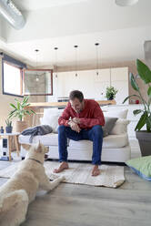 Mann sitzt auf dem Sofa vor seinem Hund zu Hause - VEGF05885