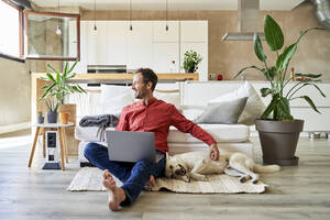 Happy businessman sitting with laptop and petting dog at home - VEGF05892