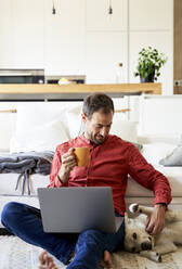 Freelancer sitting by dog holding coffee cup at home - VEGF05899