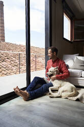 Contemplative man sitting with dog looking through window - VEGF05912