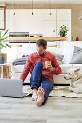 Businessman holding coffee cup working on laptop by dog - VEGF05914