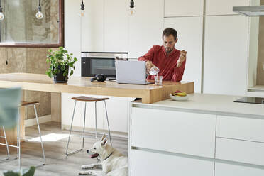 Businessman pouring water in glass sitting with laptop at home - VEGF05922