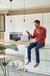 Smiling man using smart phone and eating apple at home - VEGF05923