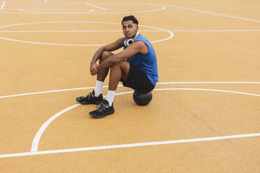 Thoughtful young man sitting on basketball at sports court - JCCMF07193