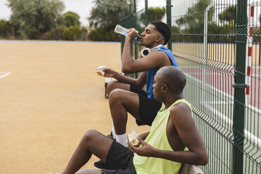 Young man drinking sports drink sitting by father on bench - JCCMF07198