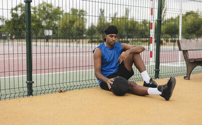 Junger Mann mit Basketball an Zaun gelehnt auf Sportplatz sitzend - JCCMF07204