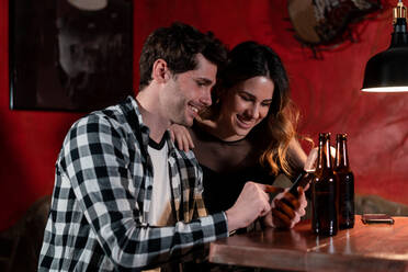 Lächelndes Paar sitzt am Tisch mit Bierflaschen und surft auf dem Handy, während es den Abend in einer Kneipe verbringt - ADSF37536