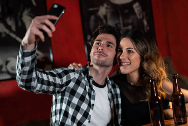 Loving couple sitting at table with bottles of beer and taking self shot on smartphone while chilling in pub at weekend - ADSF37537