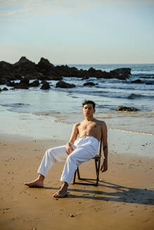 Junger ethnischer Mann in weißen Hosen sitzt und ruht sich auf einem Stuhl am nassen Strand in der Nähe des wogenden Meeres aus - ADSF37545