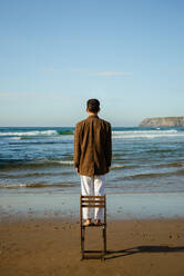 Back view of anonymous male model in stylish suit standing on chair on beach and admiring waving sea - ADSF37546
